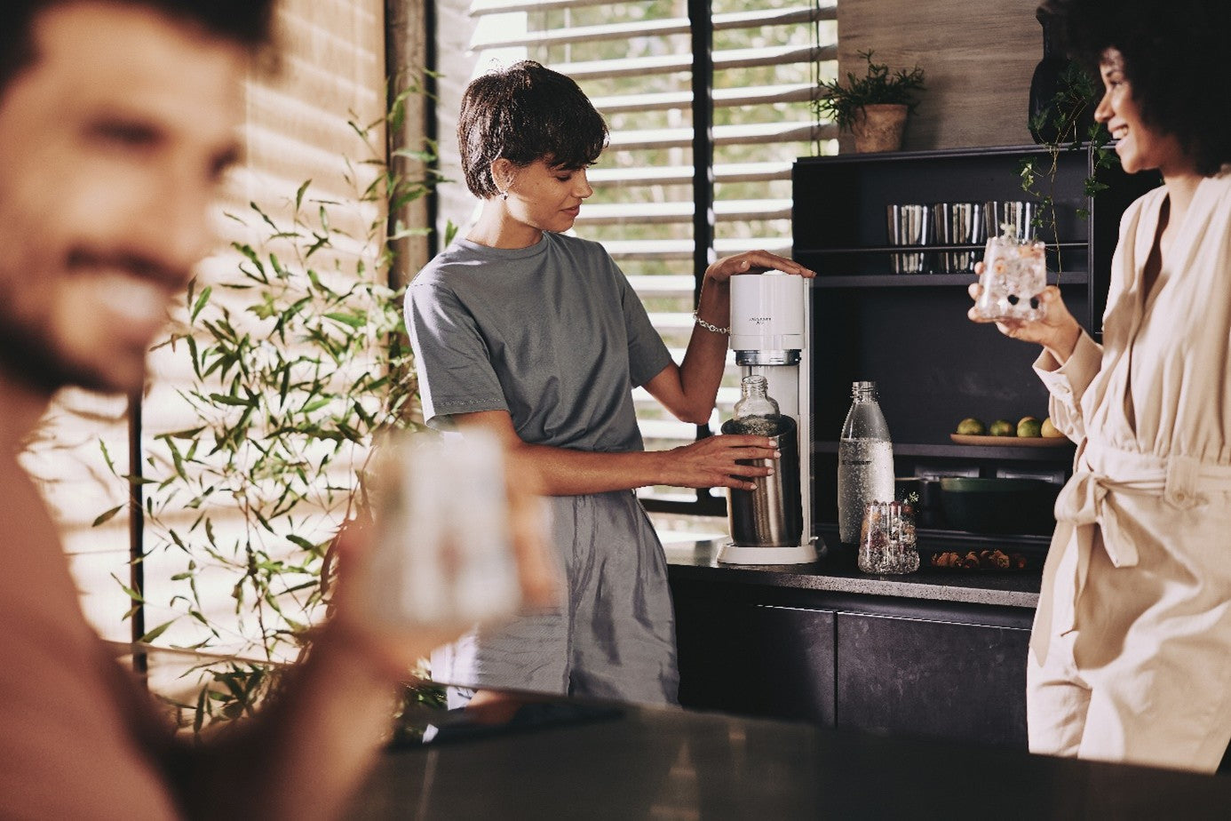 Trójka ludzi cieszy się pijąc koktajle i przygotowując wodę gazowaną w saturatorze SodaStream. 