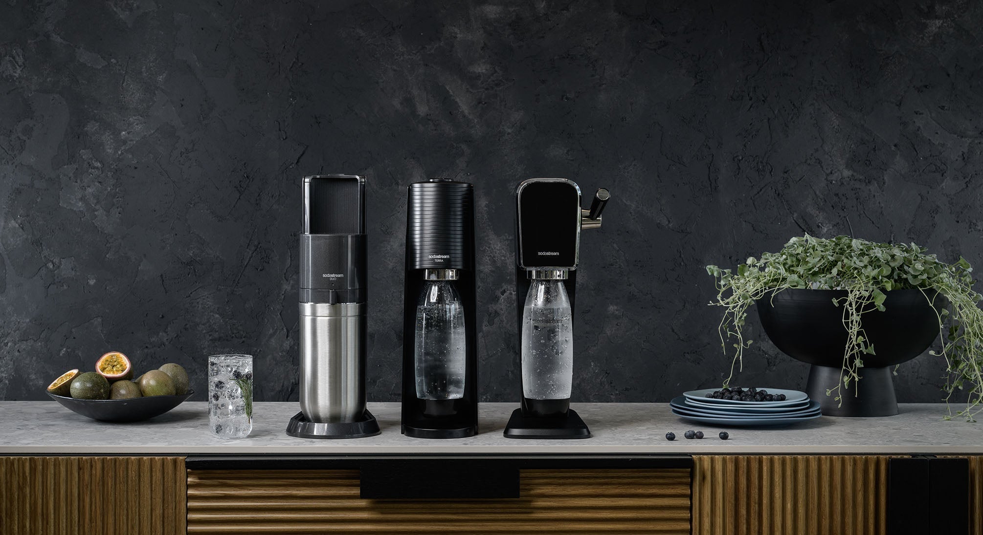 Saturatory SodaStream: Duo, Terra i Art na marmurowym blacie, obok szklanki, owoców i zielonej rośliny w eleganckiej doniczce